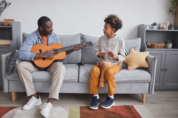 Foto completa horizontal de um homem afro-americano e seu filho passando um tempo juntos em casa tocando violão e djembê