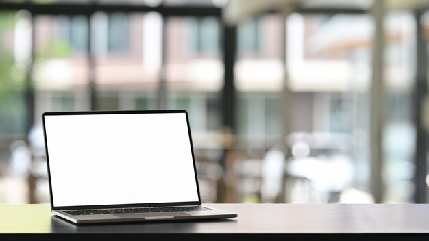 Foto de la computadora portátil con la pantalla en blanco blanca que pone en la mesa de trabajo moderna con la oficina cómoda borrosa como pantalla vacía para anunciar el concepto.