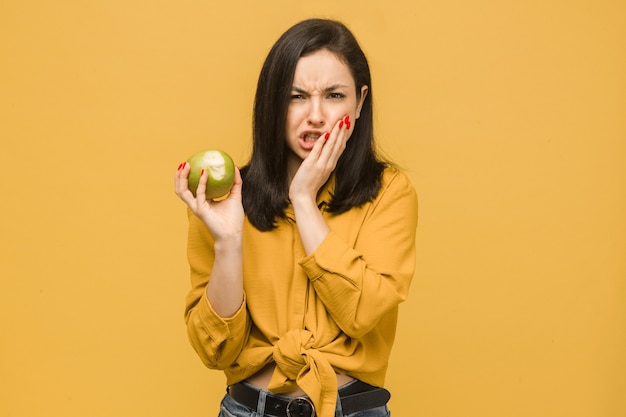 Foto de concepto de mujer joven sostiene manzana y tiene dolor de muelas. Viste camisa amarilla, fondo de color amarillo aislado.