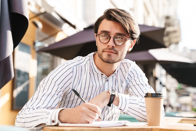 Foto de confiado hombre sin afeitar con anteojos escribiendo en el diario y tomando café mientras trabajaba en la cafetería al aire libre