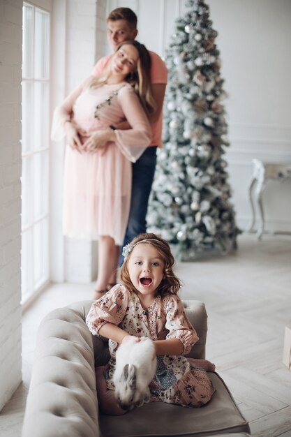 Foto conservada em estoque da linda garota brincando com o coelho no sofá. Os pais dela aninhados no fundo ao lado da árvore de Natal decorada.