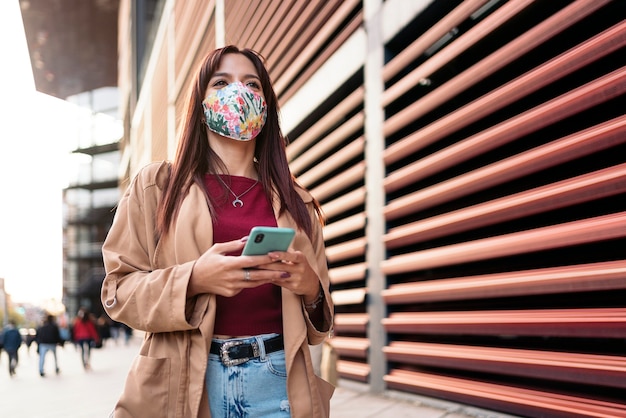 Foto conservada em estoque de uma jovem mulher caucasiana, usando seu smartphone na rua. Ela está usando uma máscara devido a covid19.
