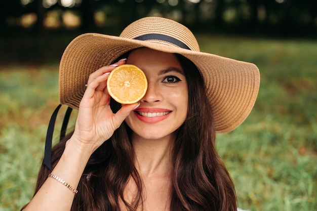 Foto conservada em estoque retrato de menina bonita com longos cabelos castanhos, com chapéu de verão elegante segurando meia laranja na mão no parque. Sorrindo para a câmera de felicidade.