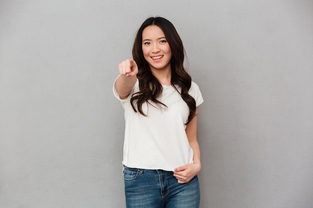 Foto de contenido mujer 20s en camiseta casual y jeans apuntando con el dedo a la cámara con una sonrisa, aislado sobre la pared gris