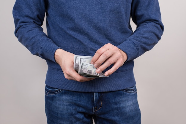 Foto cortada em close de um cara confiante em um suéter azul pulôver segurando uma pilha de dinheiro nas mãos, vestindo roupas jeans isoladas de fundo cinza