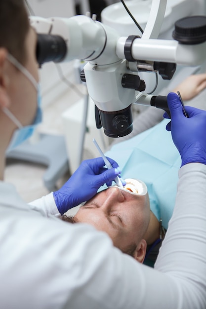 Foto cortada vertical de um paciente do sexo masculino fazendo tratamento odontológico por dentista experiente, usando um microscópio