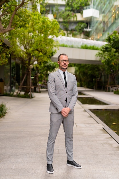 Foto de cuerpo completo del empresario hispano calvo barbudo con anteojos con la naturaleza en la ciudad