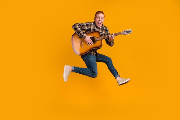 Foto de cuerpo completo de un joven saltando tocando guitarra acústica aislada sobre fondo de color amarillo