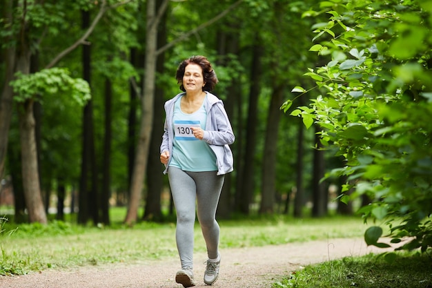 Foto de cuerpo completo de mujer madura moderna con cabello castaño corriendo maratón en el parque forestal