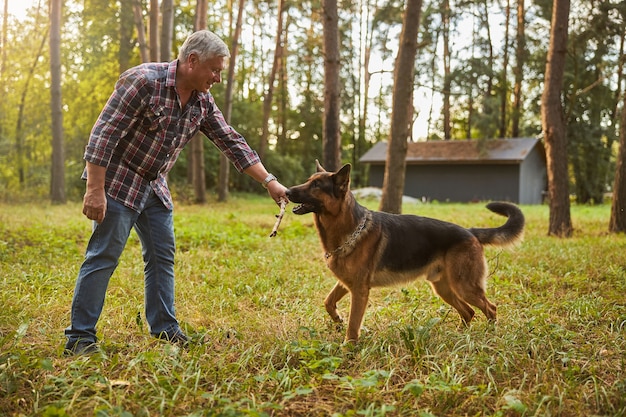 Foto de cuerpo entero de un anciano alegre dando un palo a su pastor alemán durante la reproducción