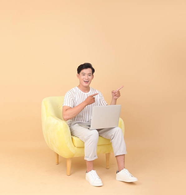 Foto de cuerpo entero de un apuesto joven asiático sentado en un sillón amarillo aislado en un fondo beige Concepto para dispositivos de alta tecnología para usuarios de aplicaciones o promoción de Internet