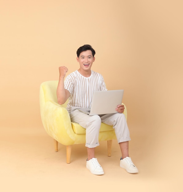 Foto de cuerpo entero de un apuesto joven asiático sentado en un sillón amarillo aislado en un fondo beige Concepto para dispositivos de alta tecnología para usuarios de aplicaciones o promoción de Internet