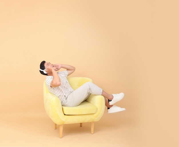 Foto de cuerpo entero de un apuesto joven asiático sentado en un sillón amarillo aislado en un fondo beige Concepto para dispositivos de alta tecnología para usuarios de aplicaciones o promoción de Internet