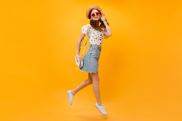 Foto de cuerpo entero de una chica con falda corta, top recortado y sombrero Mujer con gafas rojas sostiene una bolsa y corre sobre un fondo naranja