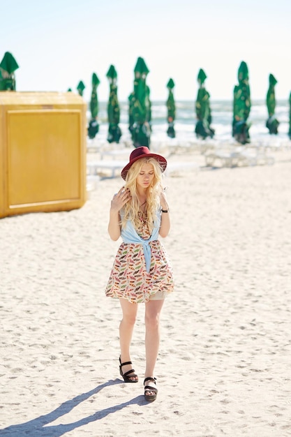 Foto de cuerpo entero de una chica rubia con el pelo rizado vestido con un vestido y un sombrero caminando en el fondo de la playa junto al mar