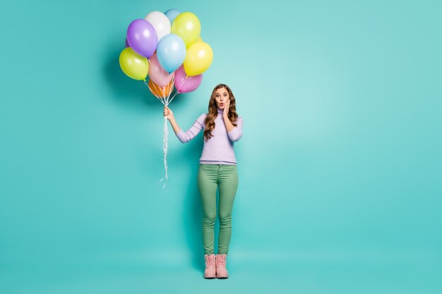 Foto de cuerpo entero de una dama bastante sorprendida llevar muchos globos de aire de colores inesperados vestidos de fiesta sorpresa lila jumper pantalones verdes botas color pastel verde azulado