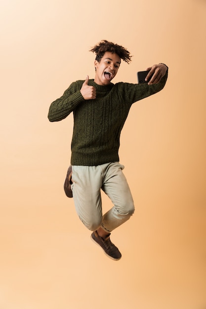 Foto de cuerpo entero de un divertido chico afroamericano con un suéter tomando selfie en un teléfono inteligente, aislado sobre una pared beige