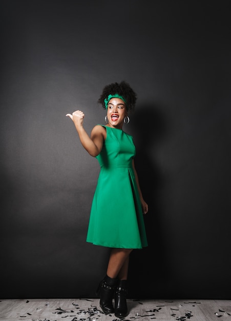 Foto de cuerpo entero de elegante dama afroamericana con vestido verde apuntando con el dedo a un lado