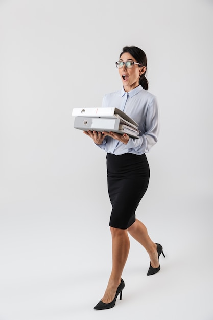 Foto de cuerpo entero de la empresaria emocionada con anteojos sosteniendo carpetas de papel en la oficina, aislada sobre la pared blanca