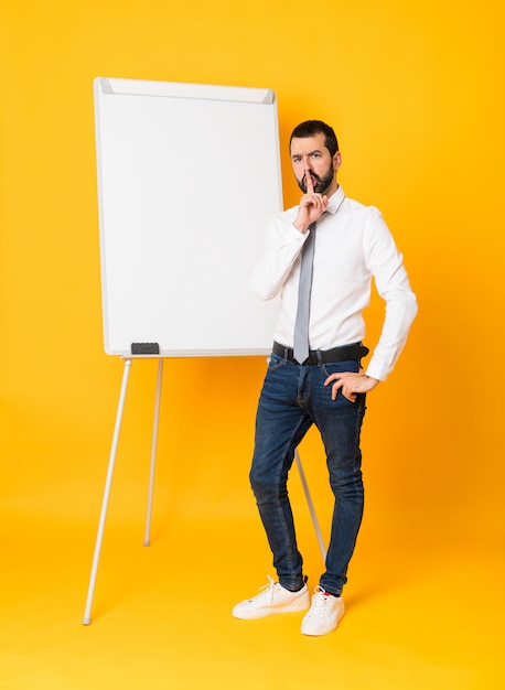 Foto de cuerpo entero del empresario dando una presentación en el pizarrón sobre amarillo aislado que muestra un signo de gesto de silencio poniendo el dedo en la boca