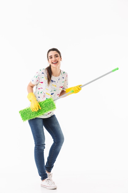 Foto de cuerpo entero de feliz ama de casa de 20 años con guantes de goma amarillos para protegerse las manos divirtiéndose mientras lava el piso con un trapeador aislado sobre una pared blanca