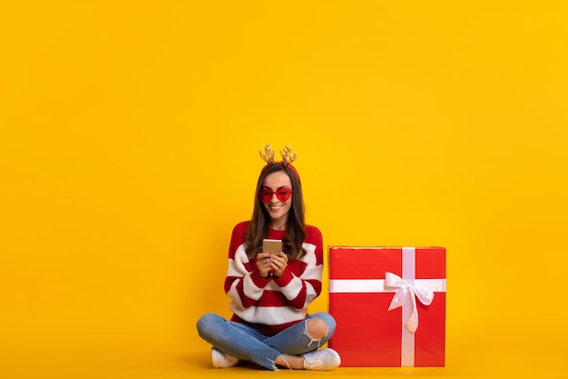 Foto de cuerpo entero de feliz joven sonriente hermosa y encantadora mujer con cuernos de reno de Navidad mientras está sentada con una gran caja de regalo roja y usando un teléfono inteligente