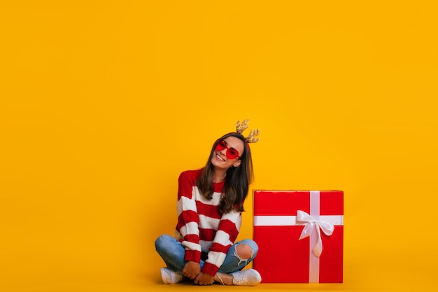 Foto de cuerpo entero de feliz joven sonriente mujer hermosa y encantadora en cuernos de reno de Navidad mientras está sentada con una gran caja de regalo roja