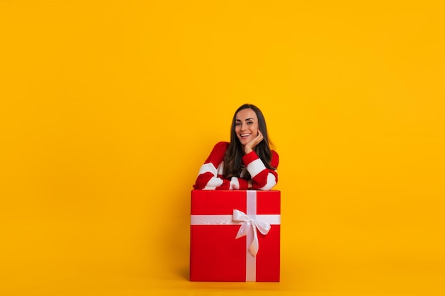 Foto de cuerpo entero de una hermosa mujer morena con estilo emocionada mientras está sentada y posando con una gran caja de regalo roja