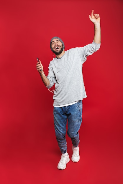 Foto foto de cuerpo entero de un hombre barbudo de 30 años cantando mientras escucha música con auriculares y teléfono móvil, aislado