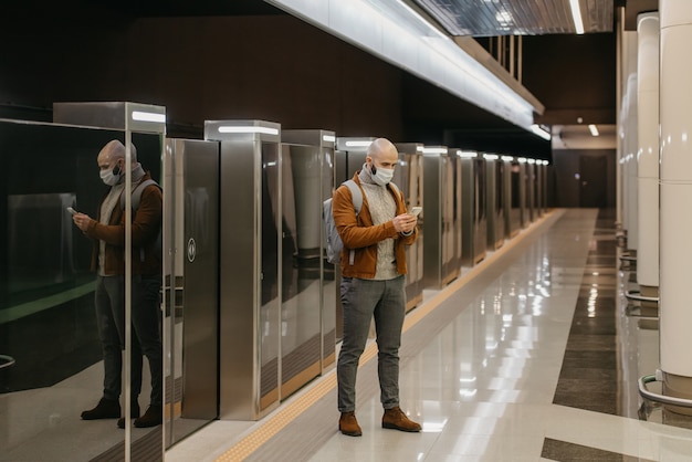 Una foto de cuerpo entero de un hombre con una máscara facial que desplaza el teléfono inteligente mientras espera un tren en la plataforma del metro. Un tipo calvo con una mascarilla quirúrgica mantiene la distancia social.