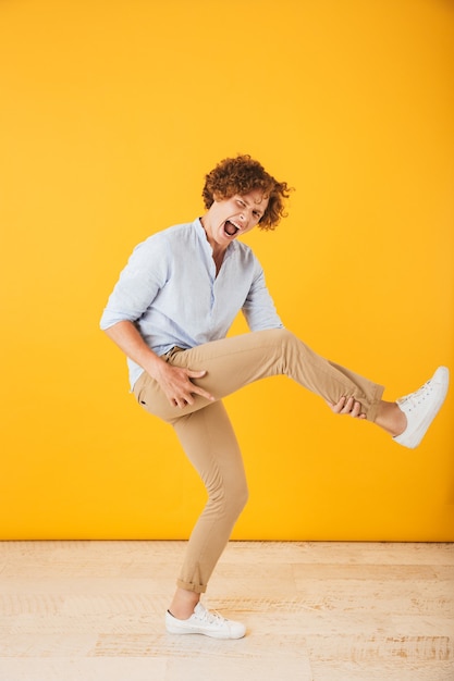 Foto de cuerpo entero de hombre rizado guapo saltando y tocando la guitarra invisible, aislado sobre fondo amarillo