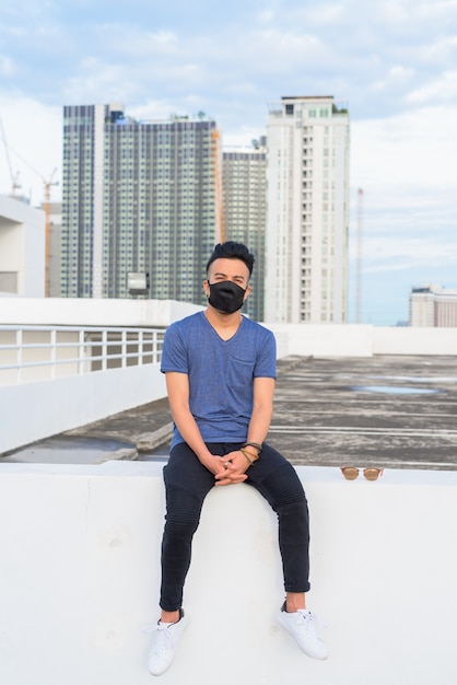Foto de cuerpo entero del joven multiétnico con máscara sentado en la azotea del edificio al aire libre