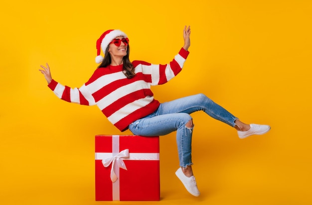 Foto de cuerpo entero de una mujer hermosa y encantadora sonriente joven emocionada con gorro de Papá Noel y con humor navideño mientras está sentada en una gran caja de regalo roja y se divierte