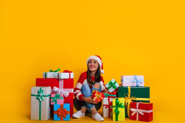 Foto de cuerpo entero de una mujer hermosa y encantadora sonriente joven emocionada con gorro de Papá Noel y con humor navideño mientras está sentada con muchas cajas de regalo coloridas y divirtiéndose