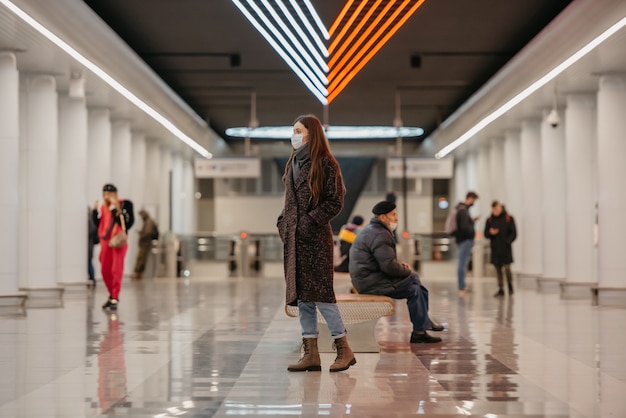 Una foto de cuerpo entero de una mujer con mascarilla para evitar la propagación del COVID que camina mientras espera un tren en el centro de una estación de metro. Chica con mascarilla quirúrgica mantiene distancia social