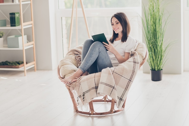 Foto de cuerpo entero de niña bonita sentarse silla de mimbre leer libro en la habitación de la casa moderna