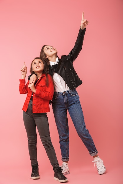 Foto de cuerpo entero de niñas sonrientes en pie casual juntas, aisladas sobre pared roja