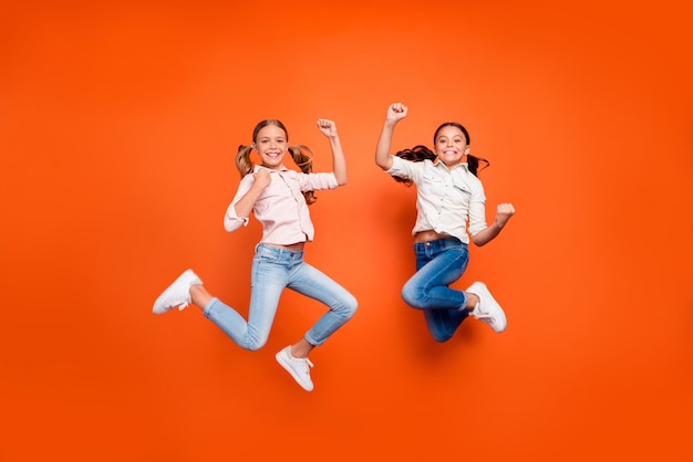 Foto de cuerpo entero de positivo lindo dos niños niñas saltar celebrar la victoria en la escuela concurso levantar puños mostrar fuerza desgaste camisa blanca pantalones vaqueros aislados fondo de color naranja