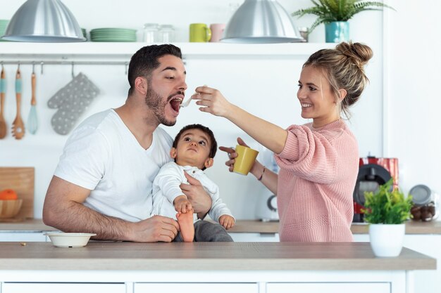 Foto foto da bela jovem mãe alimentando o marido enquanto o bebê olha para eles na cozinha em casa.