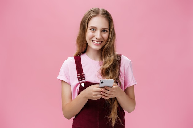 Foto da cintura da encantadora jovem agradável com cabelo louro natural ondulado em um macacão de veludo cotelê segurando o smartphone olhando para a câmera encantado e feliz discando amigo, posando sobre fundo rosa.