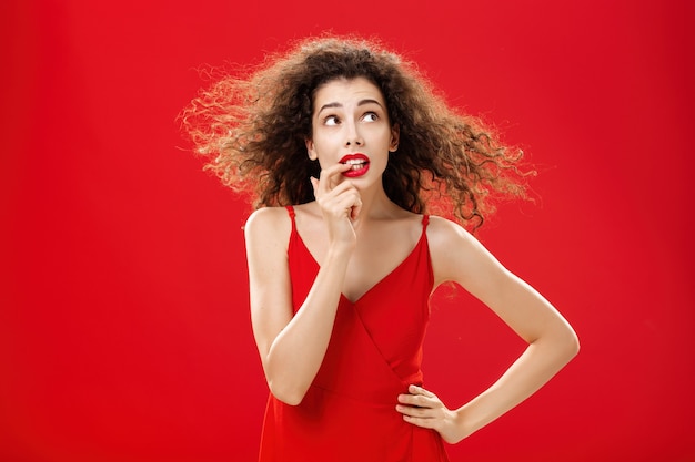 Foto da cintura de preocupada e preocupada mulher bonita insegura com penteado encaracolado em vestido de noite vermelho mordendo o dedo, parecendo incomodado ou questionado no canto superior direito com a mão na cintura. Copie o espaço