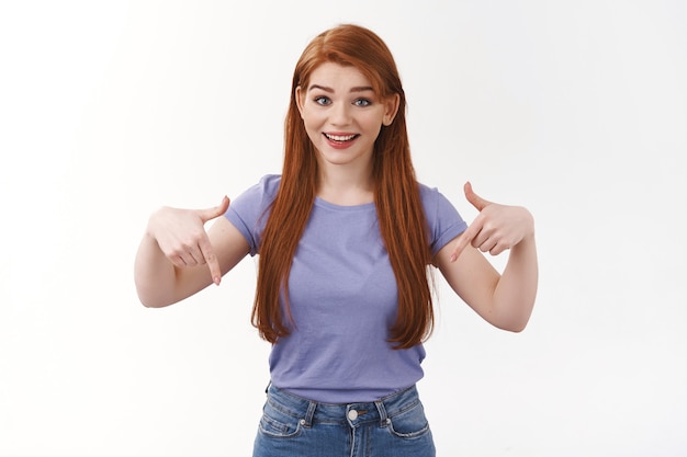 Foto da cintura para cima impressionada e animada, mulher ruiva feliz e sorridente, sorrindo em pé na parede branca em uma camiseta roxa, apontando para baixo, convide, veja, confira o banner promocional, recomende o link