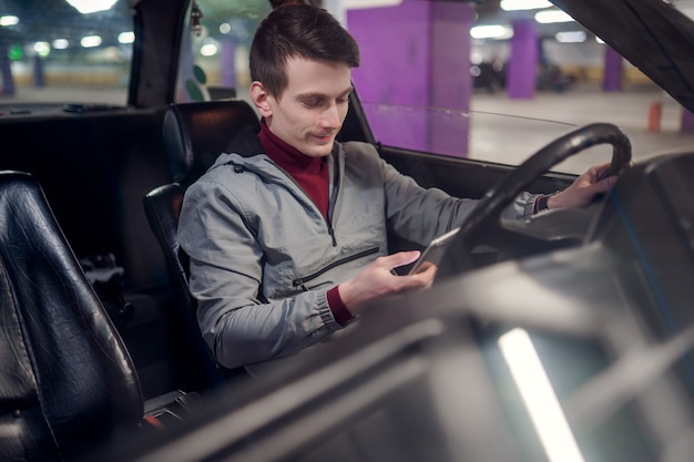 Foto da lateral de um jovem motorista do sexo masculino com o telefone nas mãos, sentado no carro