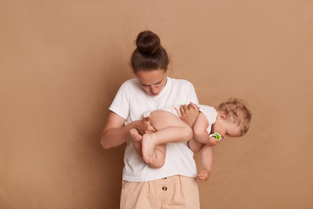 Foto da mãe olhando na fralda fedorenta de sua filhinha segurando criança infantil e faz uma careta de desgosto isolada sobre fundo marrom