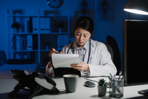 Foto da médica coreana asiática trabalhando em perícia médica enquanto está sentado na mesa na frente do computador. jovem enfermeira em roupão branco, escrevendo na área de transferência, fazendo anotações no hospital no local de trabalho escuro