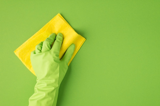 Foto da vista superior da mão na luva de borracha verde, limpando a superfície com pano amarelo sobre fundo verde isolado com copyspace à direita