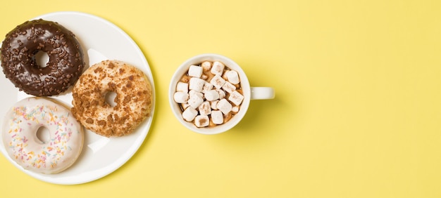 Foto da vista superior da xícara de bebida com marshmallow e prato com três rosquinhas frescas de cores diferentes em fundo amarelo pastel isolado com espaço vazio