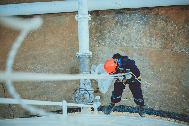 Foto da vista superior de soldador de acesso por corda industrial trabalhando em altura usando arnês, inspeção de acesso por corda de equipamento de segurança de capacete da indústria de tanques de armazenamento de espessura.