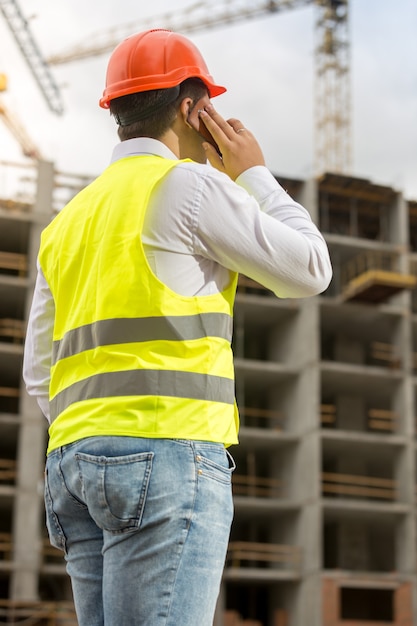 Foto da vista traseira do empresário com capacete de segurança e colete de segurança falando por telefone no canteiro de obras