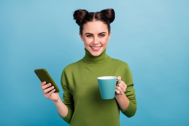 Foto de una dama adolescente bastante atractiva sostener una taza de bebida caliente disfrutar de la mañana trabajar como autónomo navegando por teléfono blog usar suéter de cuello alto verde cálido aislado pared de color azul